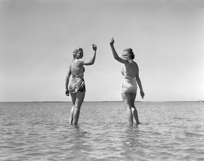 1930S Two Girls Standing In Water Wearing Bathing Suits Looking Back Waving