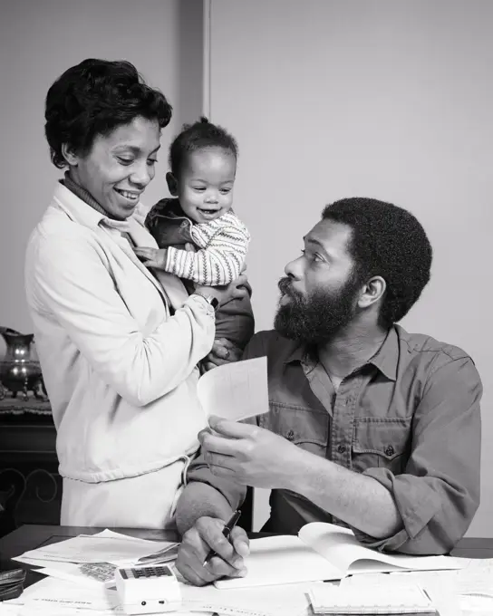 1970s SMILING AFRICAN-AMERICAN FAMILY MOTHER BABY IN ARMS TALKING WITH FATHER SITTING AT TABLE REVIEWING FAMILY BILLS AND BUDGET