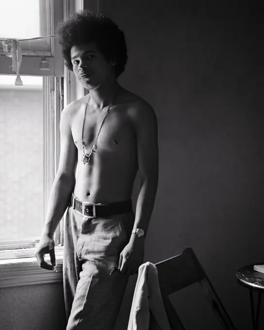 1960s IMPOVERISHED YOUNG SHIRTLESS AFRICAN-AMERICAN MAN STANDING BY WINDOW SMOKING LOOKING AT CAMERA ANGRY FACIAL EXPRESSION 