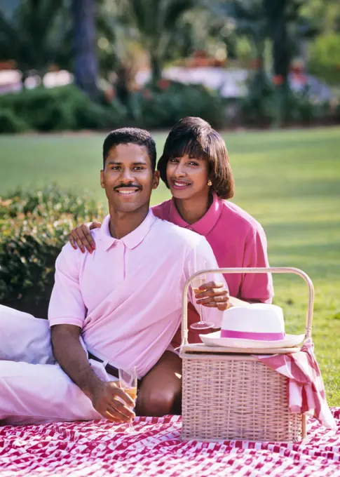 1980s 1990s AFRICAN-AMERICAN COUPLE SITTING LOOKING AT CAMERA ON BLANKET WITH PICNIC BASKET 