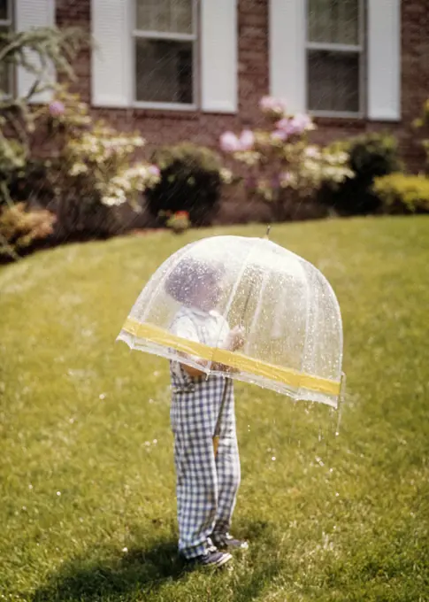 1970s 1980s LITTLE BOY UNDER PLASTIC TRNSPARENT BUBBLE UMBRELLA RHODODENDRON BRICK HOUSE SPRING RAIN SHOWER OUTDOOR GRASS 