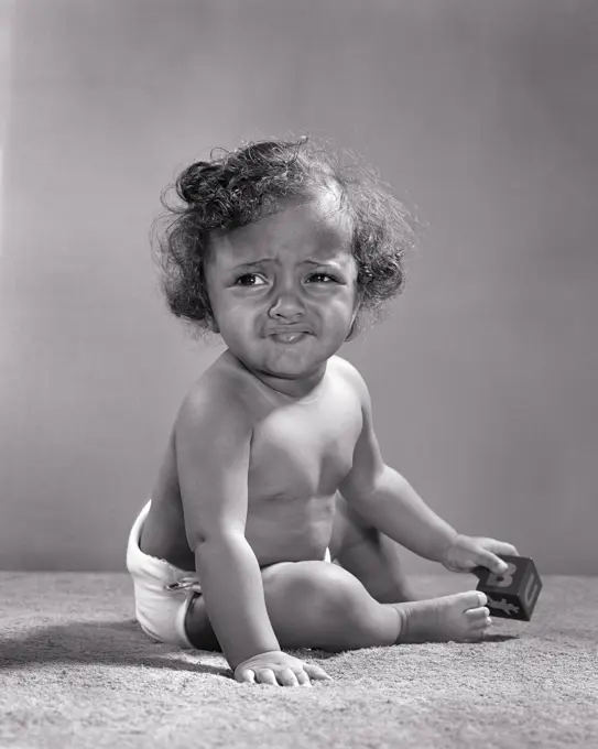 1940s 1950s UNHAPPY 1 YEAR OLD AFRICAN-AMERICAN BABY GIRL WEARING DIAPER SITTING PLAYING WITH TOY BLOCK GRIMACING LOOKING AWAY