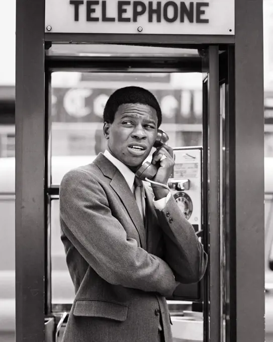 1970s ANXIOUS AFRICAN-AMERICAN MAN BUSINESSMAN AN UNSETTLED FACIAL EXPRESSION STANDING IN URBAN TELEPHONE BOOTH USING PAY PHONE