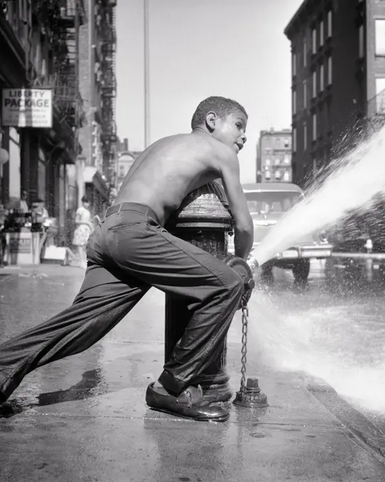 1970s ANONYMOUS TEENAGE AFRICAN-AMERICAN YOUNG MAN OPENING FIRE HYDRANT AND CREATING COOLING WATER SPRAY NEW YORK CITY NY USA