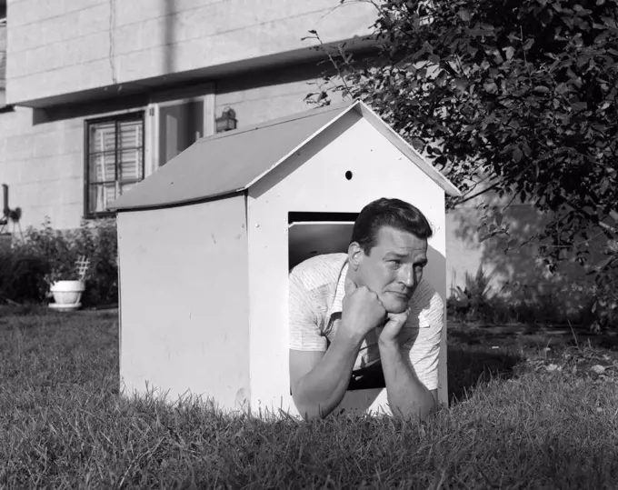 1960S Man In Doghouse In Backyard Resting On Elbows With Frown On Face