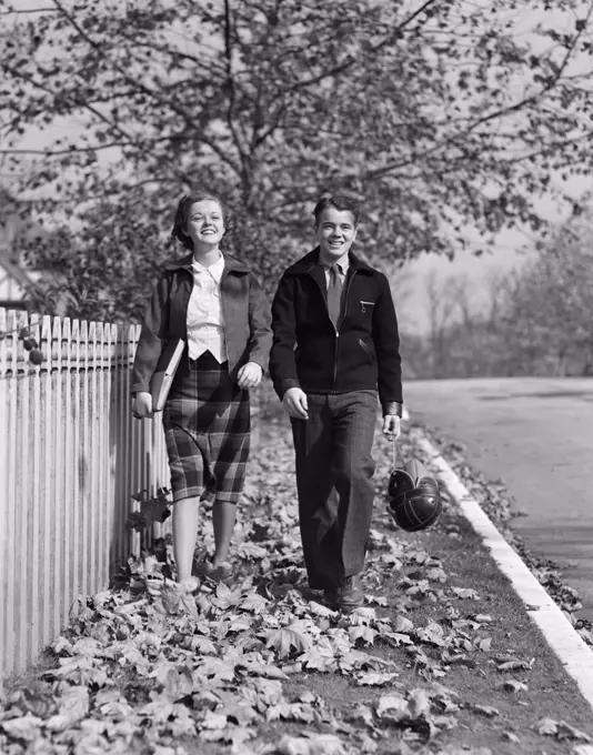 1930S Teen Couple Walking On Sidewalk In Fall Girl Carrying Schoolbooks Boy Carrying Football And Leather Helmet Smiling Outdoor