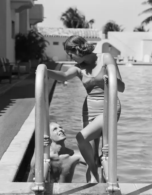 1930S Woman Wearing One Piece Bathing Suit Climbing Out Of Swimming Pool Looking Down At Man Smiling  