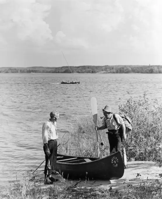 1940S 1950S Two Men Standing By River Next To Canoe One Man Holding Oar Lake Of The Woods Ontario Canada