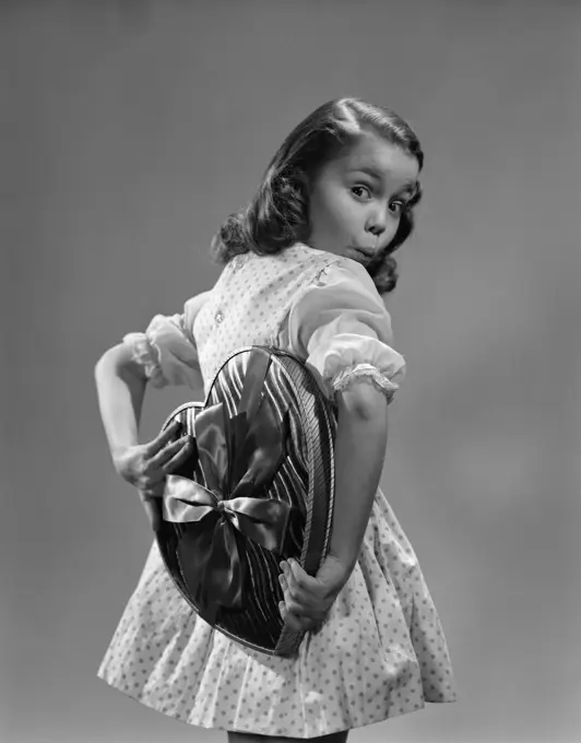 1950S Young Girl Holding Heart Shaped Candy Box Behind Back With Surprised Expression Wearing Polka Dot Dress