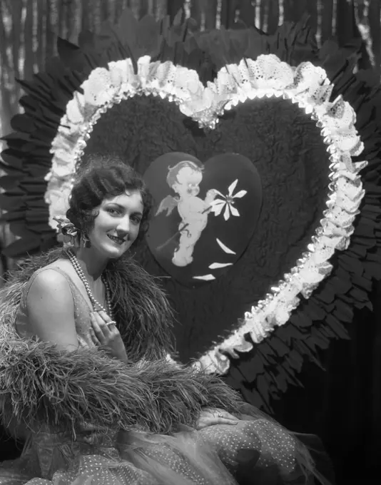 1920S 1930S Smiling Young Woman With Finger Wave Hair Pearls Feather Boa Party Dress Portrait In Front Of Giant Valentine Heart