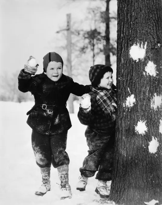 1920S 1930S Two Smiling Boys Playing In The Snow Hiding Behind Tree With Snowballs In Hand Have A Snowball Fight