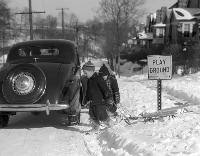 1940S Two Boys Pulling Sleds Across A Snowy Suburban Winter Street With Parked Car And Sign That Says Play Ground