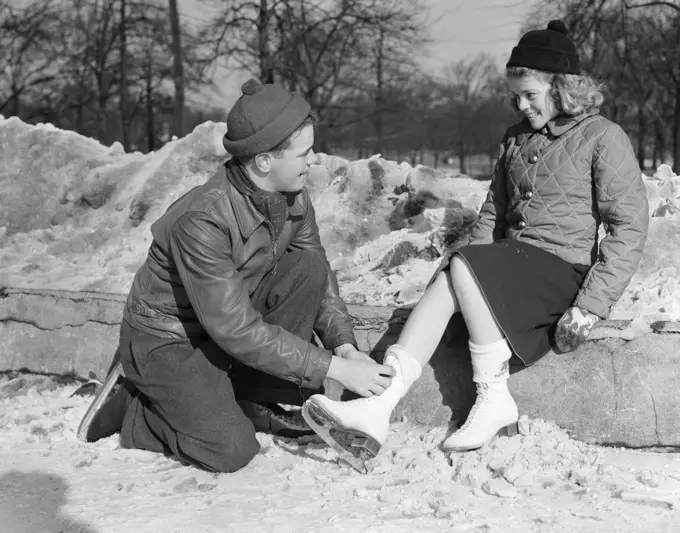 1940S Smiling Romantic Innocent Wholesome Teenage Couple In Knit Caps & Winter Jackets Kneeling Boy Tying Ice Skates For Girl