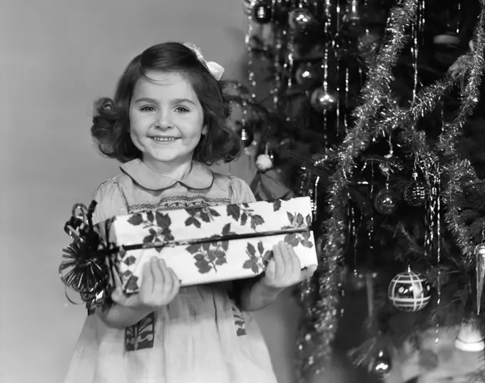 1940S Little Girl Standing Near Christmas Tree Holding A Wrapped Present Smiling