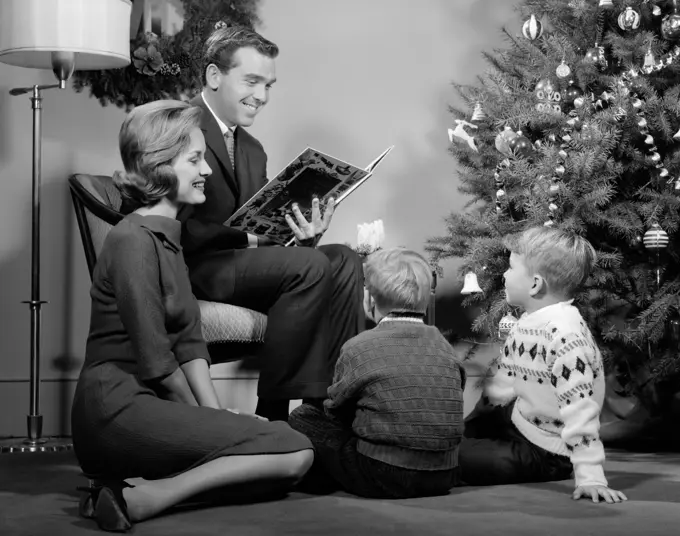 1960S Family Father Mother Two Sons Smiling Sitting By Christmas Tree In Living Room Reading A Book