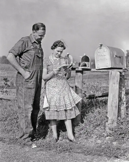 1930s 1940s MAN AND WOMAN FARMER AND WIFE TOGETHER COLLECTING MAIL FROM RURAL POSTAL MAILBOX READING MAGAZINE SEED CATALOG