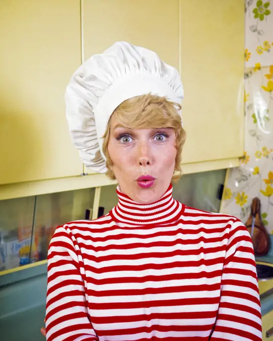 1970s BLONDE HOUSEWIFE WEARING CHEFS HAT RED AND WHITE TURTLE NECK TOP EYES WIDE OPEN MAKING FUNNY FACIAL EXPRESSION IN KITCHEN