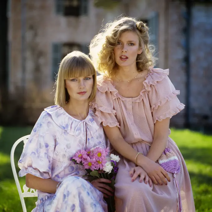 1970s 1980s TWO BLONDE WOMEN WEARING RUFFLED DRESSES IN PASTEL COLORS SITTING OUTDOORS LOOKING AT CAMERA 