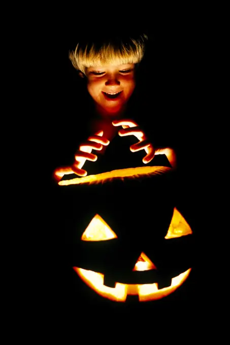 1980s BLONDE BOY IN SCARY POSE MAKING A THREATENING HAND GESTURE ABOVE AN ILLUMINATED HALLOWEEN CARVED PUMPKIN AT NIGHT