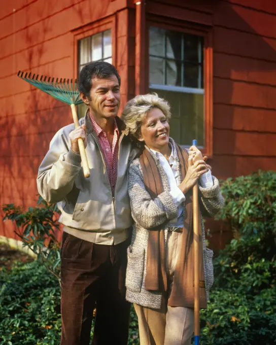 1980s MATURE COUPLE STANDING BY HOUSE MAN HOLDING RAKE ON SHOULDER WOMAN LEANING ON GARDEN TOOL WEARING AUTUMN CLOTHES SMILING