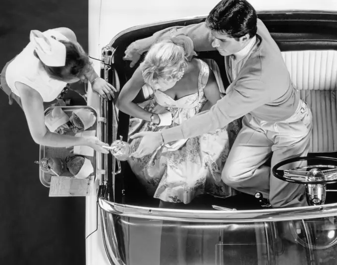 1950s DRIVE-IN RESTAURANT CARHOP WAITRESS ON ROLLER SKATES SERVING COUPLE IN 1955 T-BIRD CAR BURGERS AND MILKSHAKES