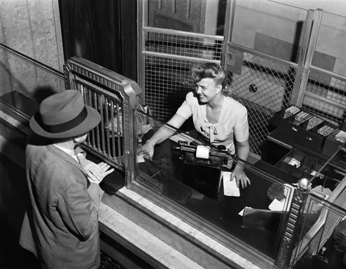 1940s WOMAN BANK TELLER BEHIND CAGE SERVING MALE CUSTOMER