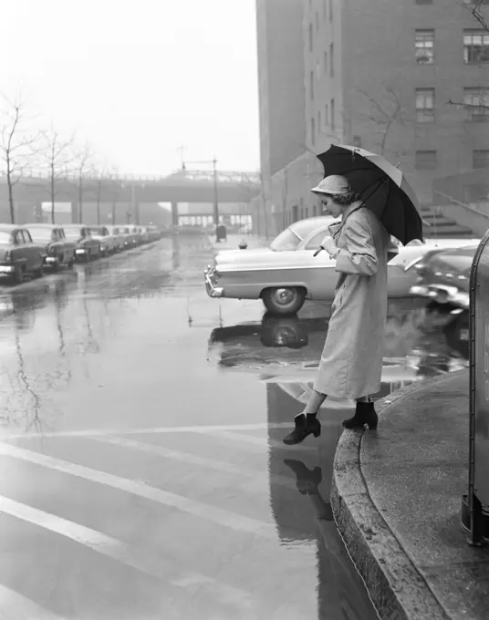 1950s WOMAN IN RAIN COAT HAT BOOTS HOLDING  UMBRELLA CROSSING CITY STREET IN WET FOUL WEATHER