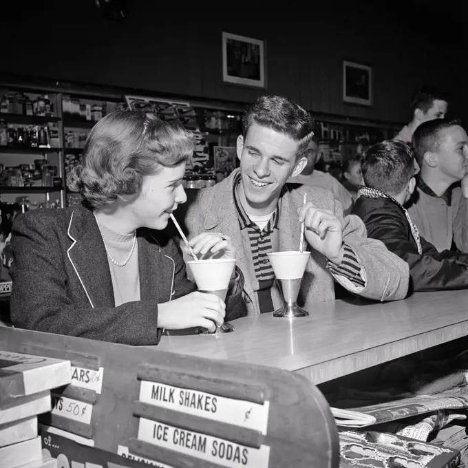 1950s TEENAGE COUPLE BOY AND GIRL SITTING TOGETHER DRINKING SOFT DRINKS AT SODA FOUNTAIN COUNTER LAUGHING SMILING FLIRTING