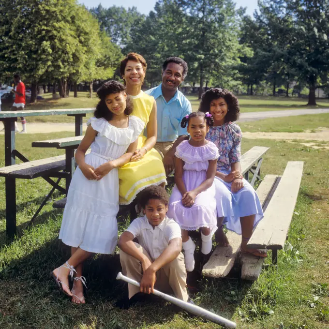 1980s PORTRAIT AFRICAN-AMERICAN FAMILY OF 6 SITTING ON PARK SPECTATOR BENCH BOY HOLDING ALUMINUM BASEBALL BAT LOOKING AT CAMERA