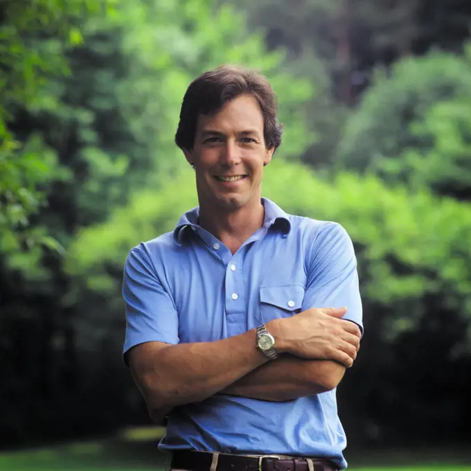 1980s SMILING PORTRAIT OF HANDSOME MAN STANDING ALONE OUTDOORS ARMS CROSSED WEARING BLUE SHORT SLEEVE SHIRT LOOKING AT CAMERA