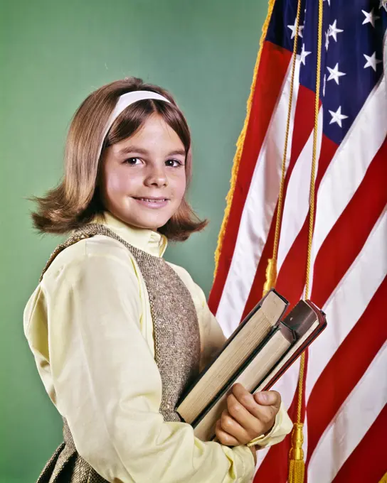 1960s SMILING GIRL WEARING TWEED JUMPER YELLOW BLOUSE HOLDING TEXTBOOKS SMILING LOOKING AT CAMERA STANDING BESIDE AMERICAN FLAG