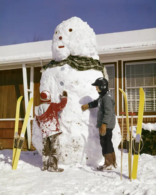1960s TWO BOYS WITH VERY TALL SNOWMAN BY HOUSE YELLOW SKIS STUCK IN SNOW