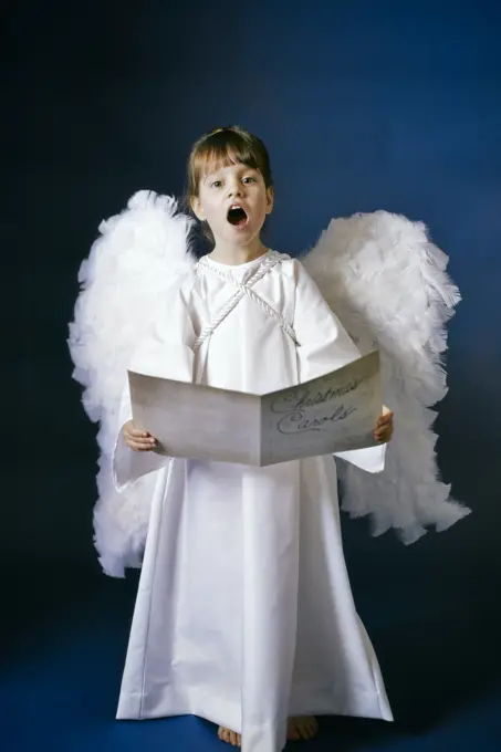 1960s CUTE LITTLE GIRL IN WHITE ANGEL GOWN COSTUME WITH WINGS LOOKING AT CAMERA SINGING CHRISTMAS CAROLS IN SUNDAY SCHOOL PLAY