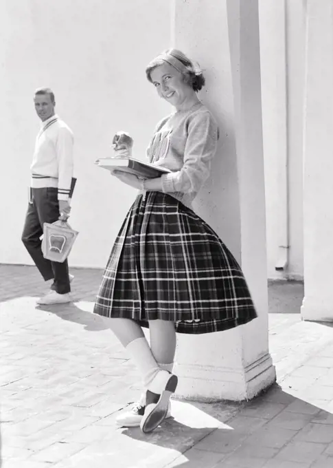 1950s 1960s TEEN GIRL LEANING AGAINST COLUMN EATING APPLE READING BOOK LOOKING AT CAMERA SMILING AS POTENTIAL BOYFRIEND WALKS BY