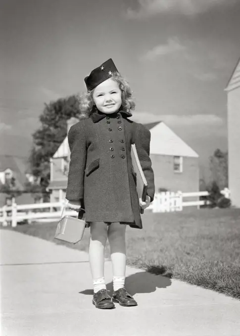 1940s SMILING LITTLE GIRL READY FOR SCHOOL STANDING ON SIDEWALK WEARING BLACK HAT DOUBLE BREASTED COAT HOLDING LUNCH BOX & BOOK