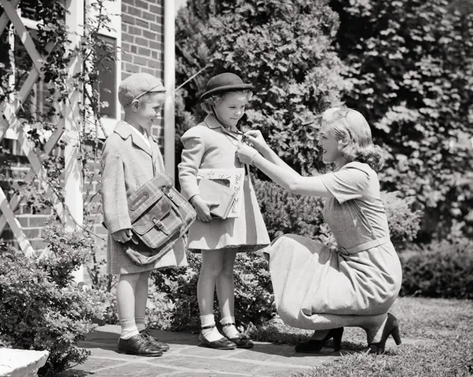 1940s 1950s MOTHER ADJUSTING CHECKING COAT ON GIRL AND BOY DAUGHTER SON SISTER BROTHER READY TO LEAVE FOR SCHOOL BOOK BAG BOOKS