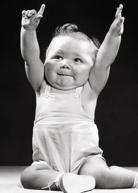 1950s BABY BOY TODDLER SITTING WITH ARMS RAISED HANDS UP ONE HAND MAKING ENTHUSIASTIC NUMBER ONE SIGN