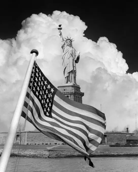 1940s COMPOSITE IMAGE OF STATUE OF LIBERTY WITH DRAMATIC CLOUD BEHIND AND 48 STAR AMERICAN FLAG OVERLAY IN FOREGROUND