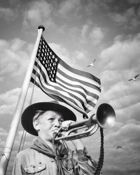 1930s 1940s BOY SCOUT IN UNIFORM BLOWING A BUGLE IN FRONT OF A 48 STAR AMERICAN FLAG 