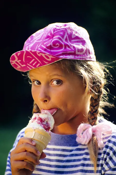 1980s WIDE-EYED GIRL WITH BRAIDS EATING ICE CREAM CONE WEARING PINK BALL CAP LOOKING AT CAMERA