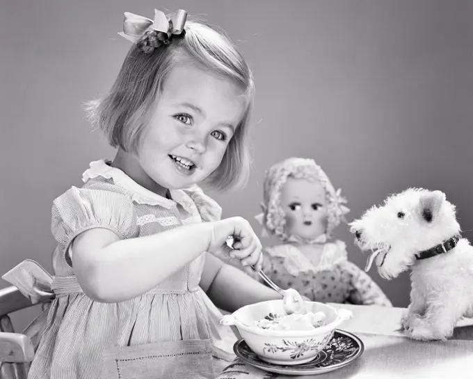 1940s 1950s SMILING GIRL BOW IN HAIR EATING BOWL OF ICE CREAM LOOKING AT CAMERA SURROUNDED BY STUFFED ANIMAL DOG AND DOLL TOYS 