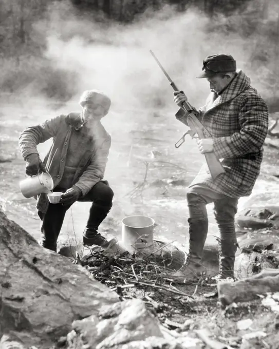 1920s TWO HUNTERS STANDING CAMPED BY STREAM ONE MAKING COFFEE OVER CAMPFIRE ONE CHECKING LEVER ACTION RIFLE NEW BRUNSWICK CANADA