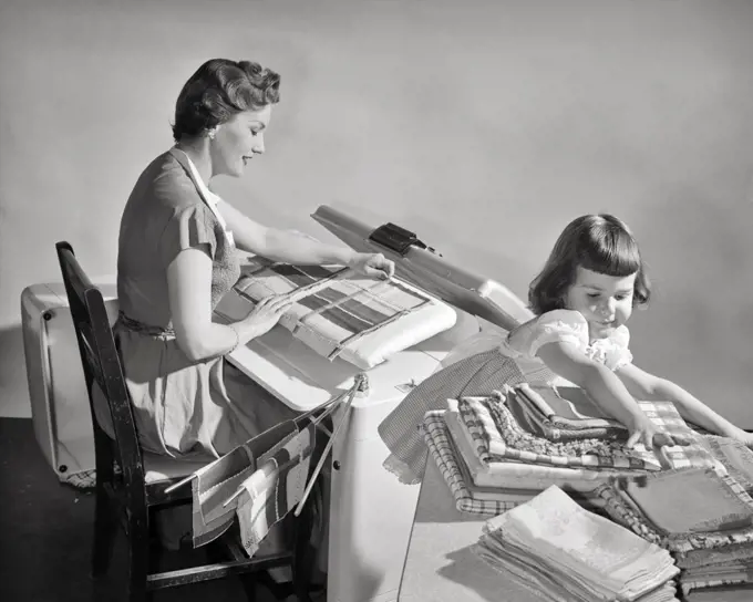 1950s WOMAN HOUSEWIFE USING AN ELECTRIC APPLIANCE STEAM PRESS TO IRON LINENS AS GIRL DAUGHTER FOLDS AND STACKS THEM ON TABLE