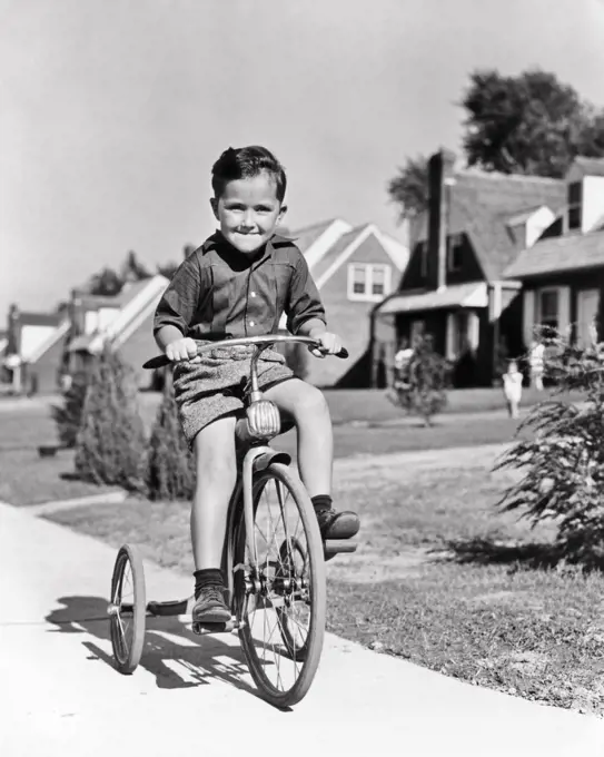 1930s 1940s BOY RIDING TRICYCLE BIKE ON SUBURBAN SIDEWALK SMILING HAPPY DETERMINED FACIAL EXPRESSION LOOKING AT CAMERA 