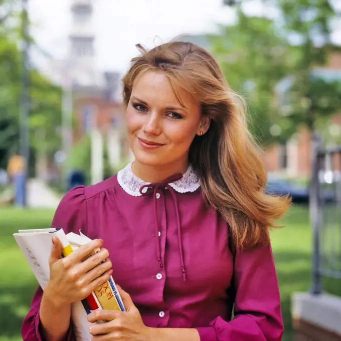 1980s PRETTY SMILING DISHWATER BLONDE COED GIRL STUDENT ON CAMPUS HOLDING TEXT BOOKS WEARING MAROON BLOUSE LOOKING AT CAMERA 