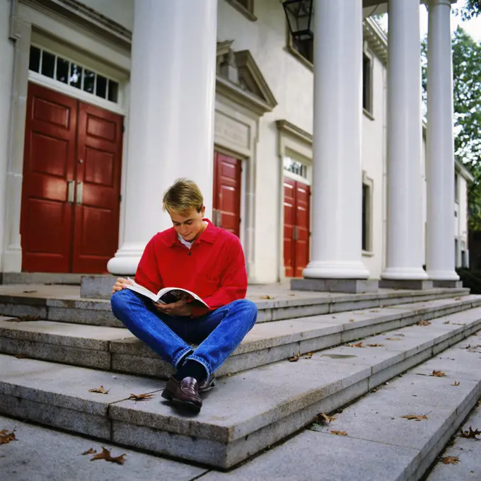 1980s MALE COLLEGE STUDENT RED SHIRT BLUE JEANS SITTING ON SCHOOL BUILDING STEPS STUDYING READING TEXTBOOK 