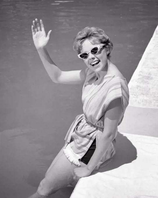 1960s smiling YOUNG woman sitting on edge of swimming pool waving greeting wearing sunglasses