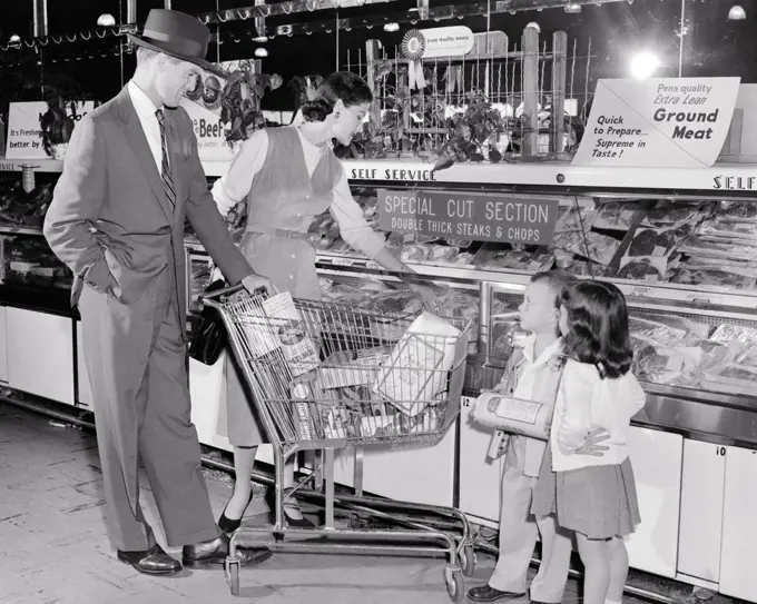 1950s FAMILY OF FOUR SHOPPING IN GROCERY STORE WITH CART AT MEAT COUNTER MOTHER FATHER WITH SON AND DAUGHTER