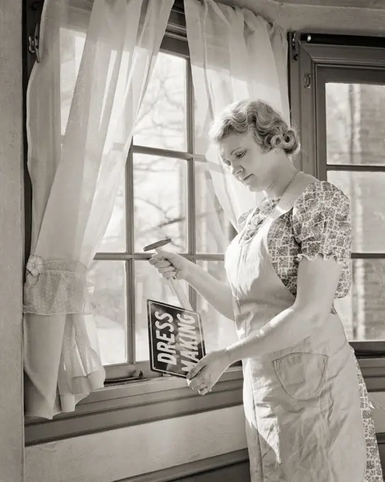 1940s MIDDLE AGED WOMAN PUTTING A DRESS MAKING SERVICES SIGN IN THE WINDOW OF HER HOME