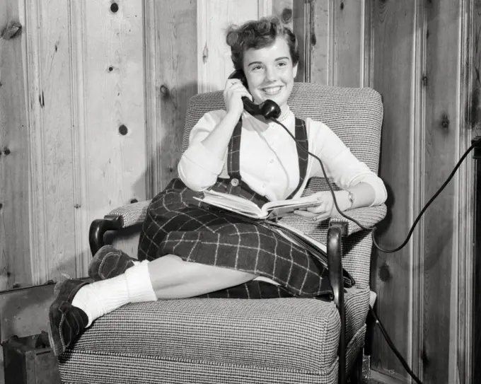 1950s TEEN GIRL SMILING TALKING ON THE TELEPHONE READING BOOK IN HER LAP WEARING BOBBY SOCKS LOAFERS SITTING IN EASY CHAIR